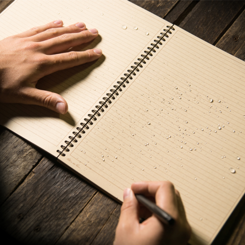 close up of person writing in spiral waterproof notebook on a wooden table