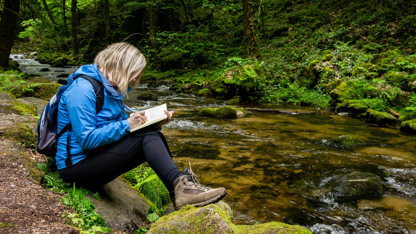 woman writing in a notebook sat by a river outdoors